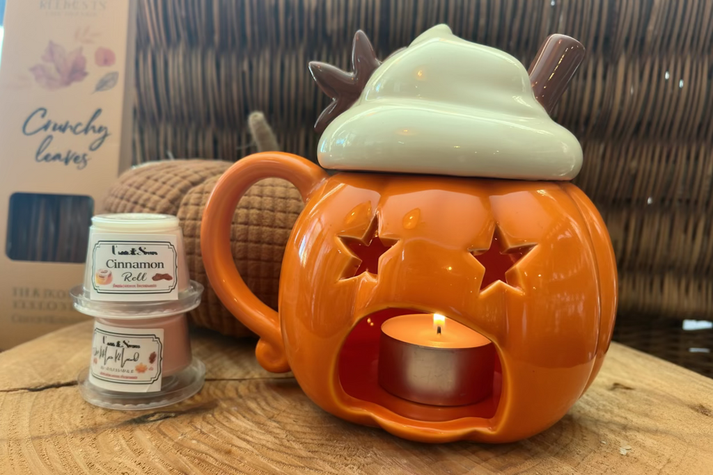 Chauffe-cire citrouille orange allumé laissant filtrer la lueur d'une bougie, avec son couvercle en crème chantilly posé à côté sur une table en bois.